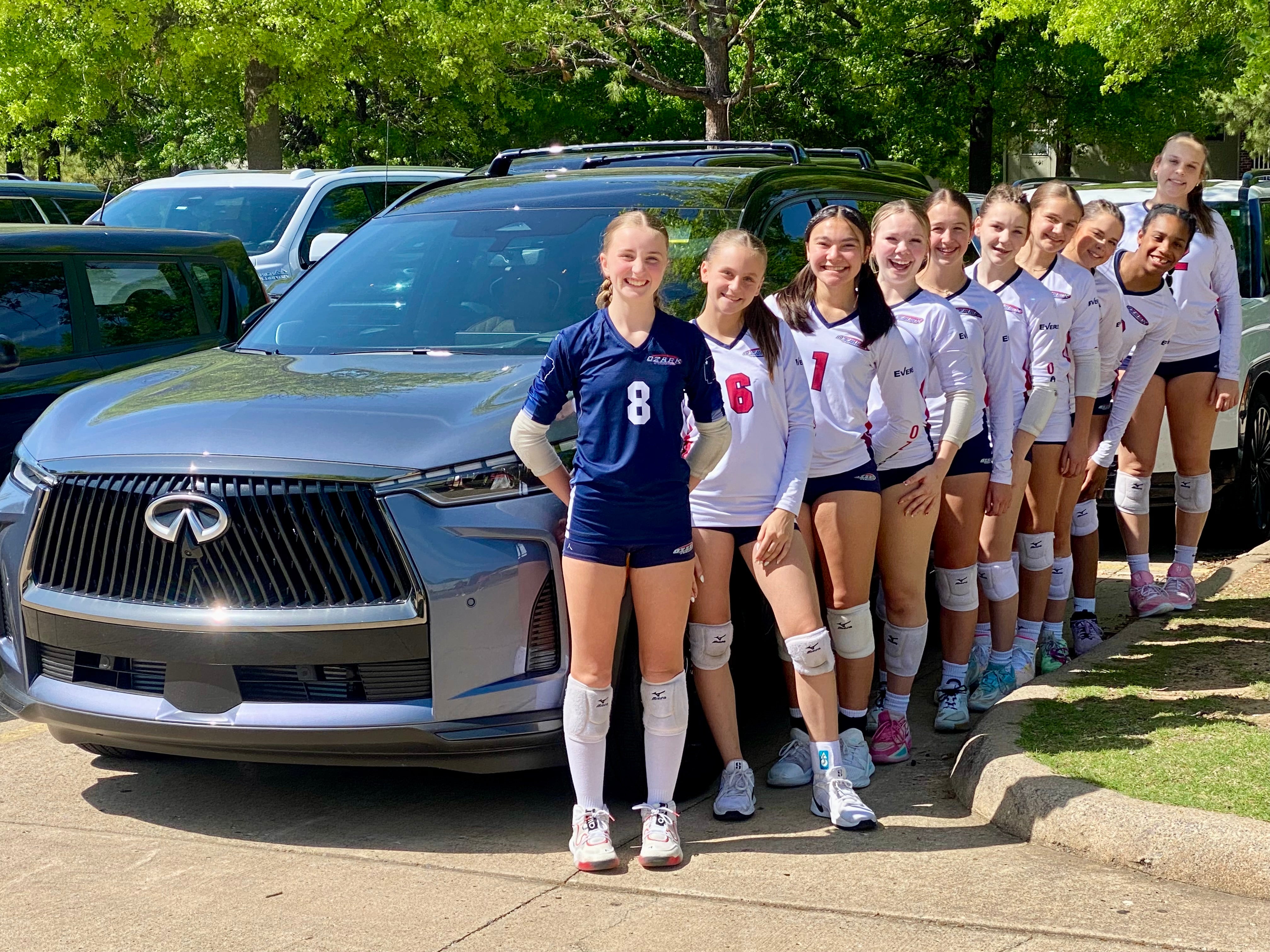 A dozen jovial female volleyball players in uniforms, knee pads, and athletic shoes, standing by a dark-gray SUV.