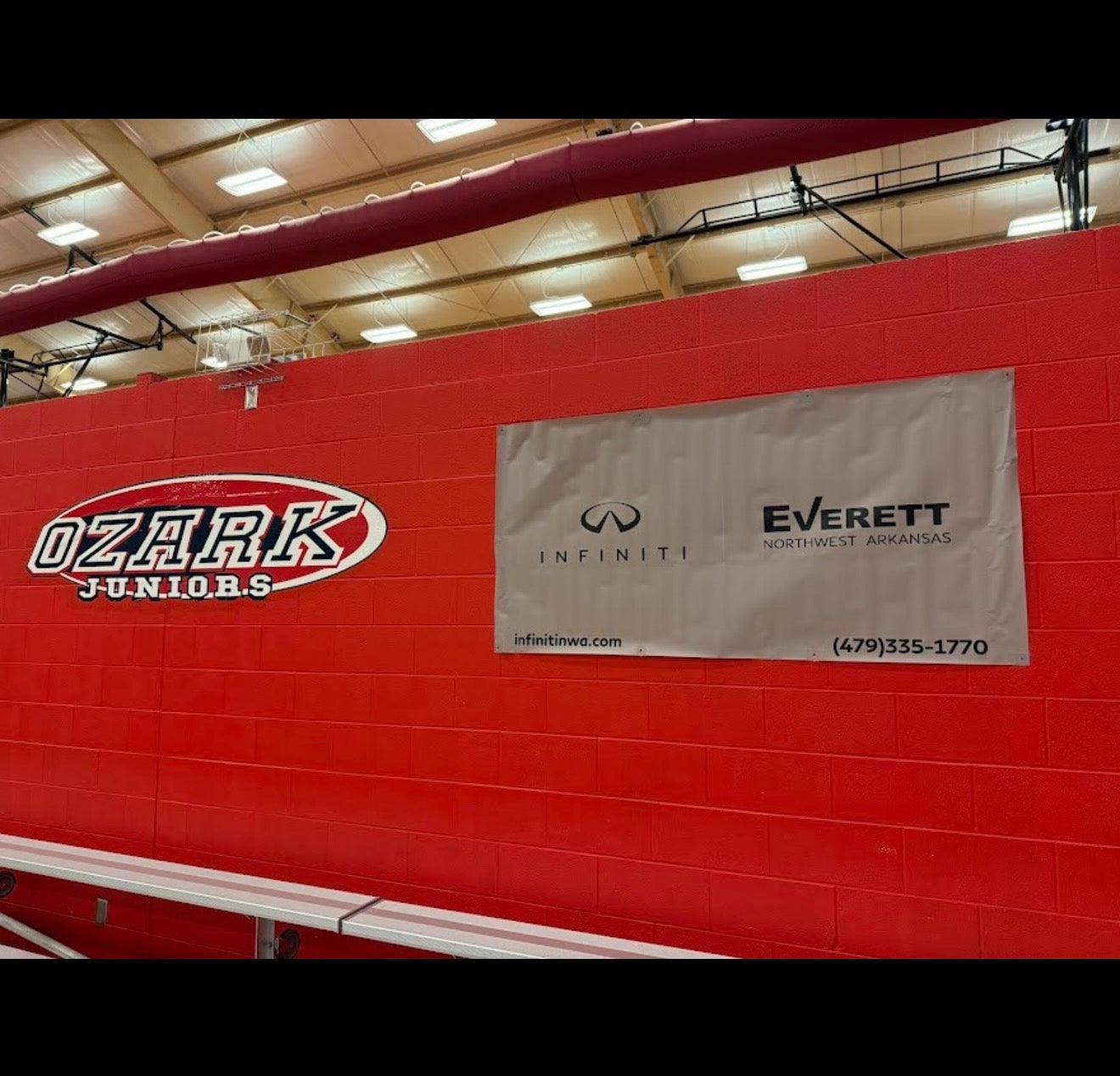 Ozark Juniors logo and Infiniti dealership banner on a vibrant red brick wall in a gymnasium.