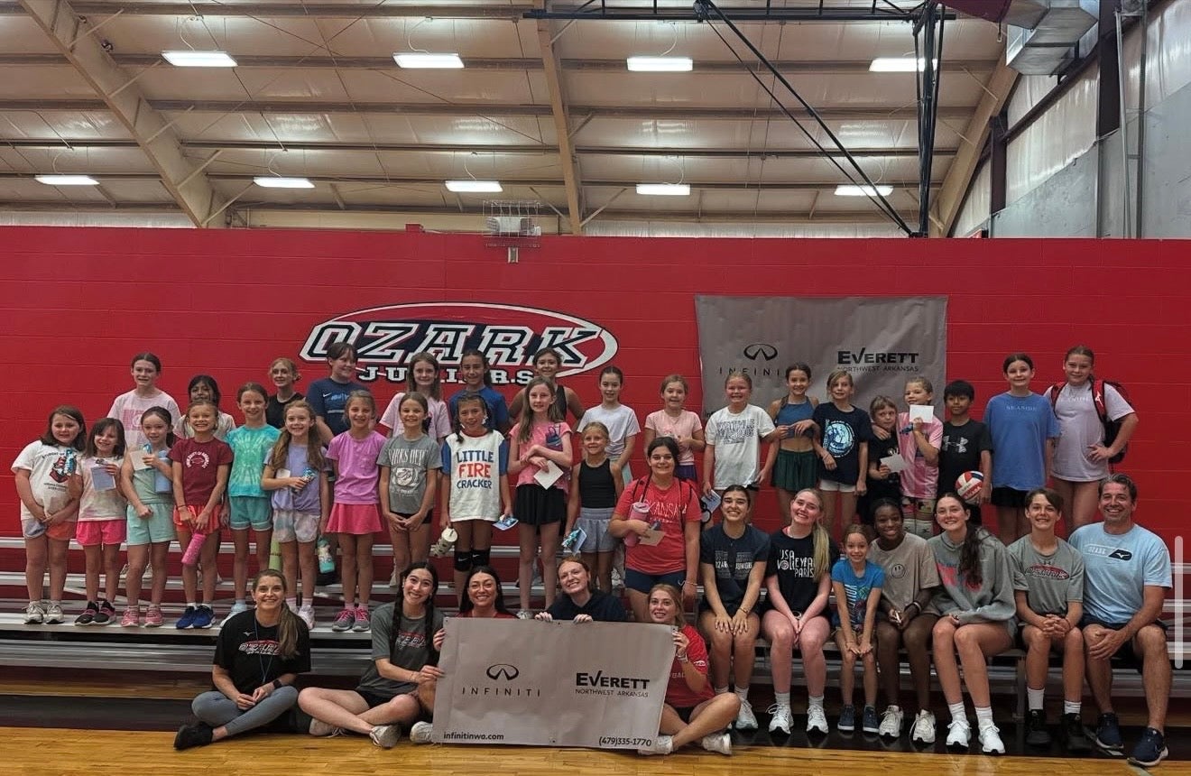 A large group of young volleyball players and coaches pose with a sponsor banner in a gymnasium.