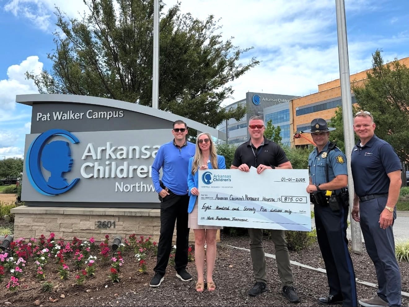 Five people, including a police officer, stand by the Arkansas Children's sign, holding a large check for $875.