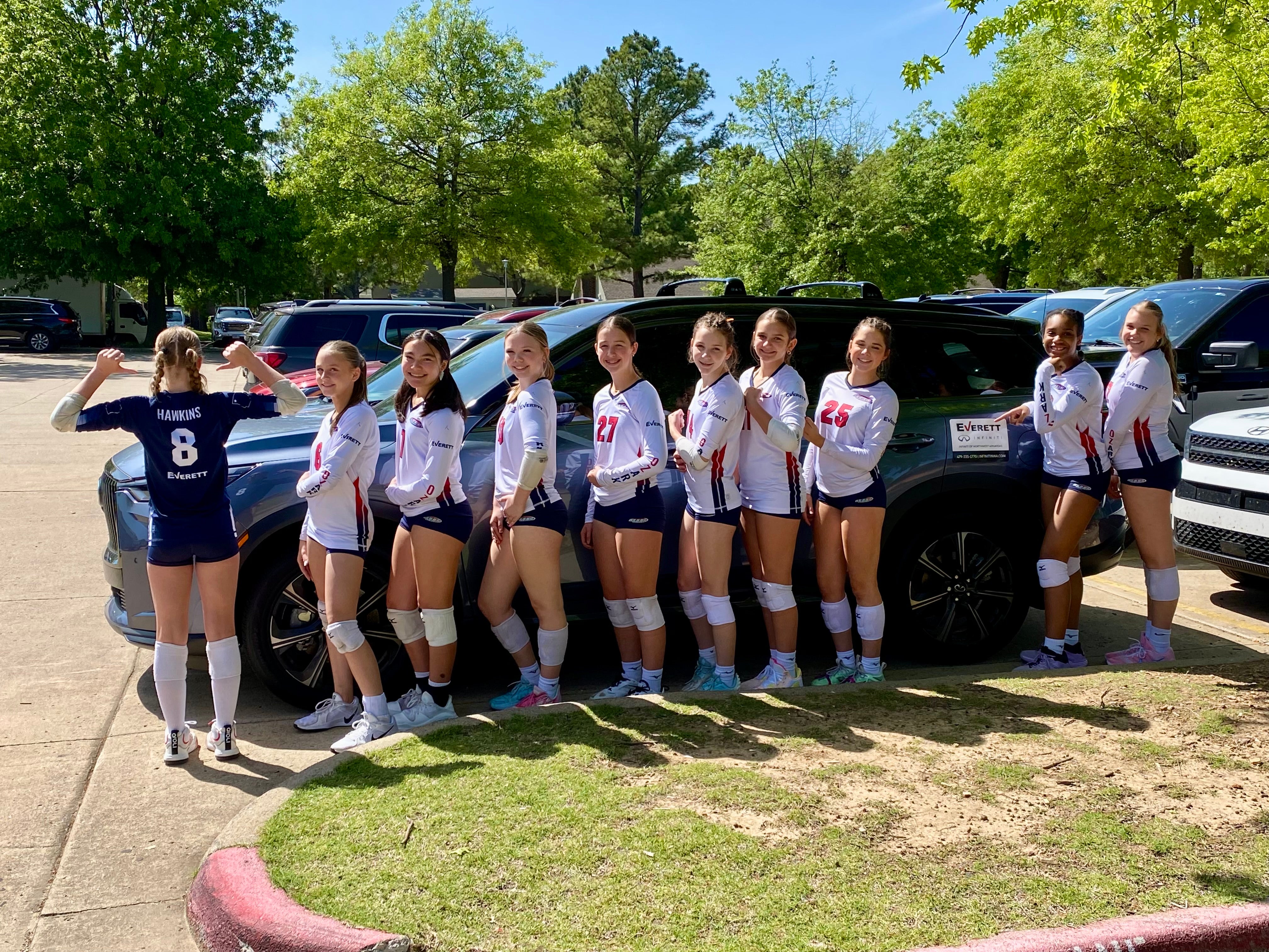 A volleyball team of eleven smiling girls in uniforms posing in a line in a parking lot.