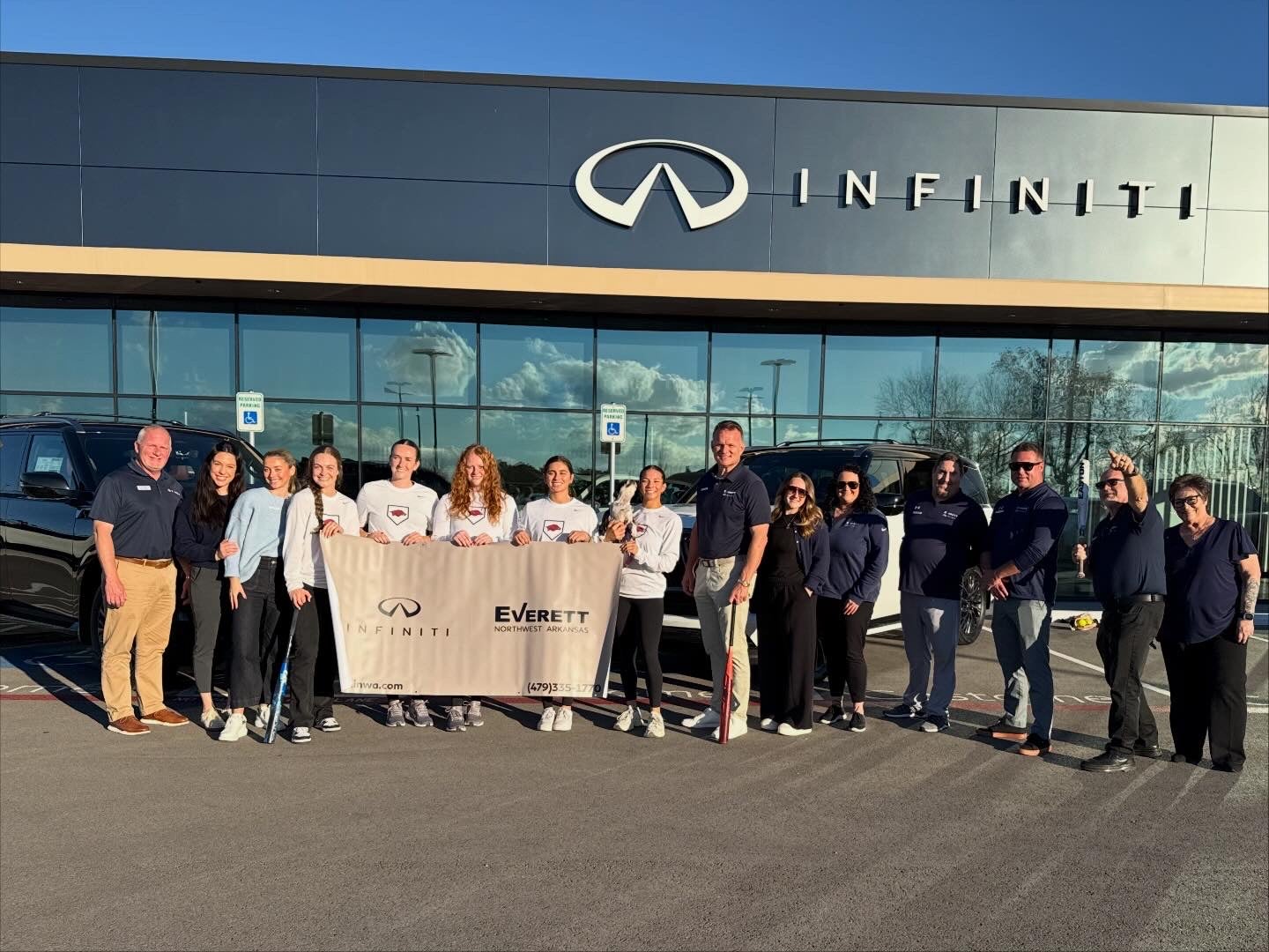 A group of 15 adults, including 6 female athletes with lacrosse sticks, holding a banner in front of an Infiniti dealership.