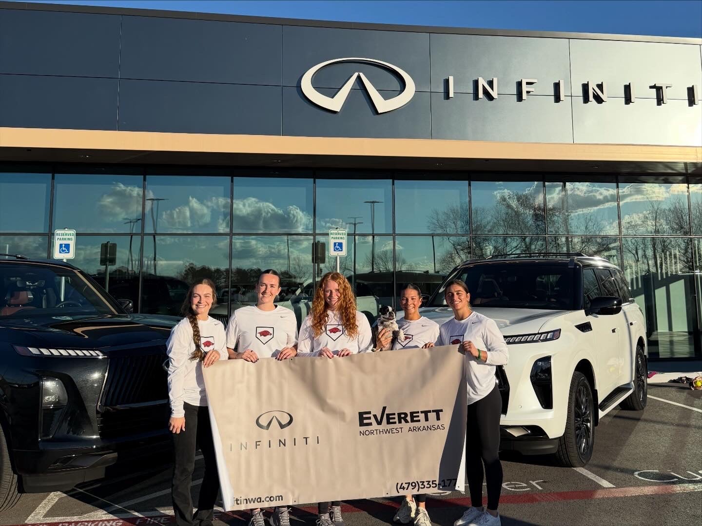 Five young women, one holding a small dog, stand outside an Infiniti dealership holding a banner.