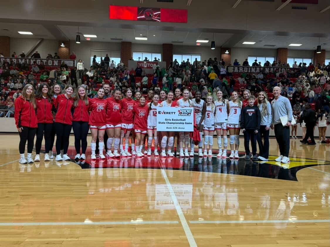 A girls' basketball team in red and white jerseys, holding a championship game banner on a basketball court.