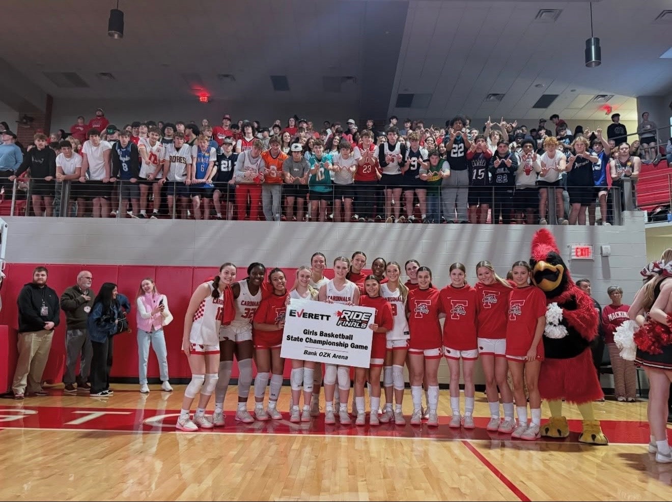 Girls' basketball team in white and red uniforms posing with a State Championship Game sign and red mascot.