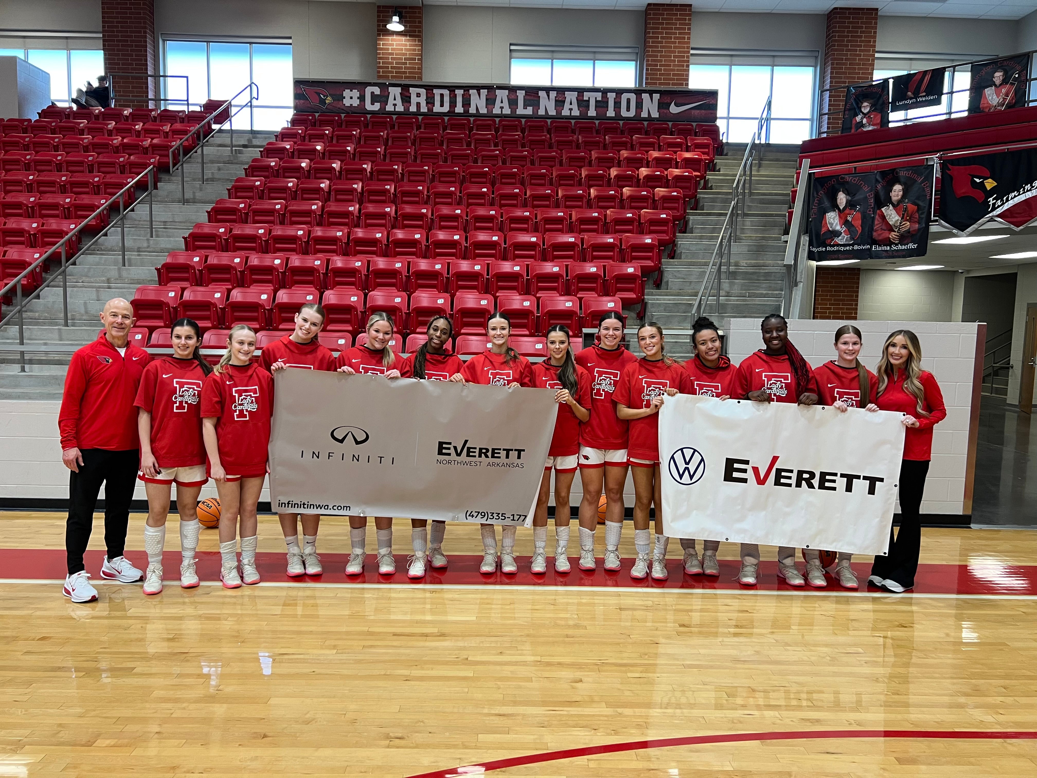 A basketball coach, a woman, and 12 female basketball players in red shirts holding two sponsor banners.